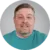 A man with short brown hair and a goatee, wearing a teal shirt, smiles slightly at the camera against a gray background.