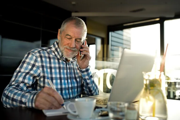 An older man in a checkered shirt talks on a phone while writing in a notebook and using a laptop at a cafÃ©. A cup of coffee and a glass container are on the table.