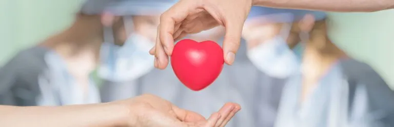 Hands holding a red heart shape, with medical personnel in scrubs and masks blurred in the background.