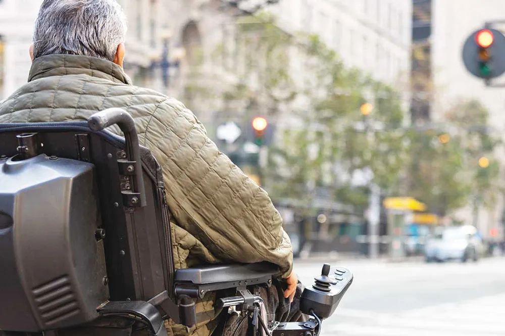 A person in a powered wheelchair crosses a city street, wearing a green quilted jacket. Traffic lights and buildings are visible in the background.