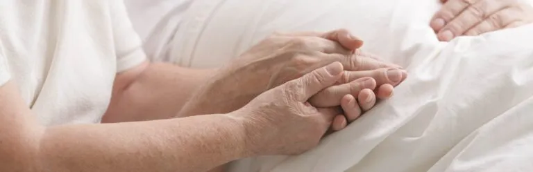Close-up of an elderly person's hands gently holding another person's hands on a white bedspread.