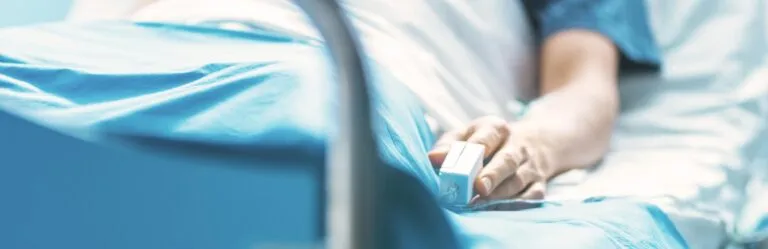 A person's hand resting on a hospital bed, wearing a pulse oximeter, signifying a medical setting where Medicare Part A covers care.