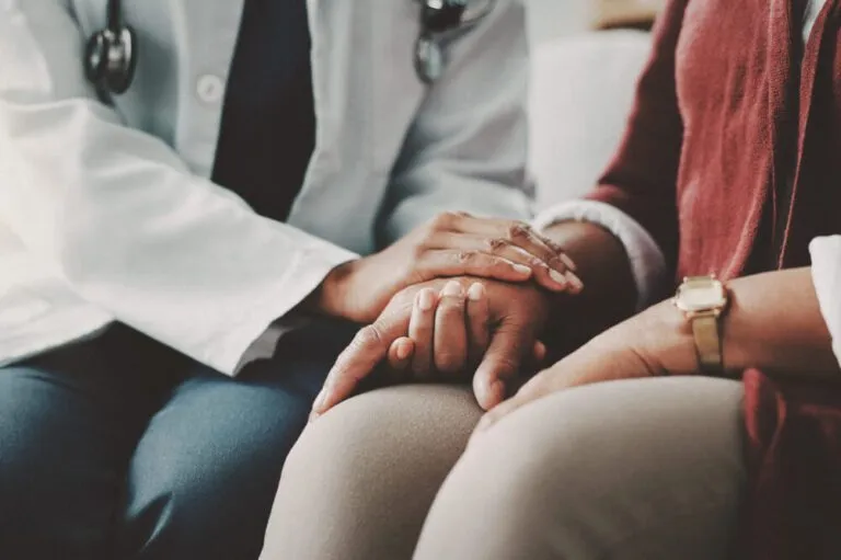 A female doctor comforting a patient by holding her hand while sitting beside her