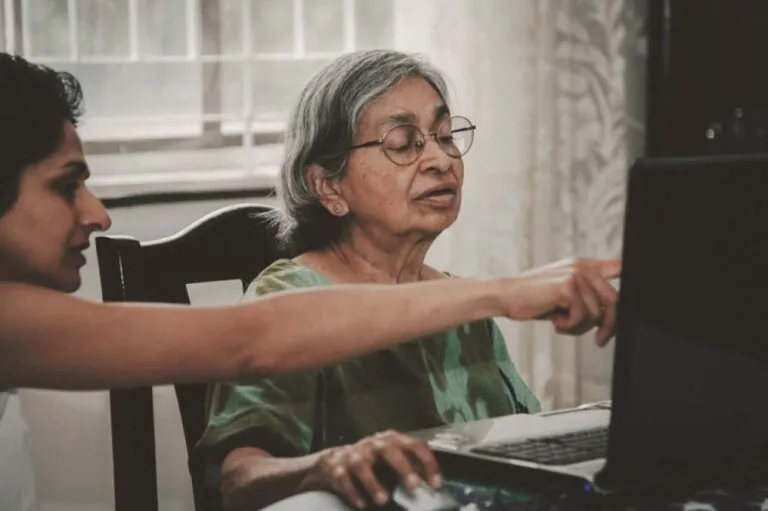 A senior mother working on a computer with assistance from her adult daughter