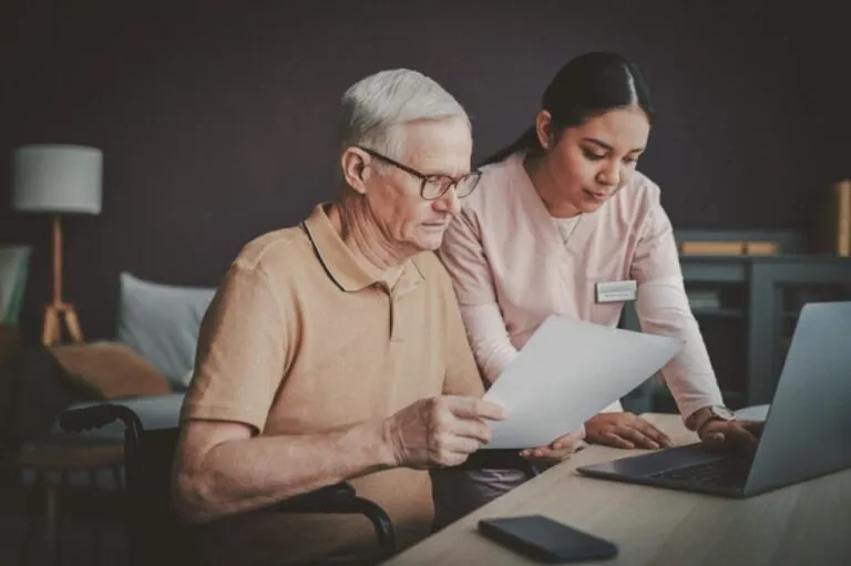 A nurse and a senior man in a wheelchair are looking at a laptop on the desk in front of them