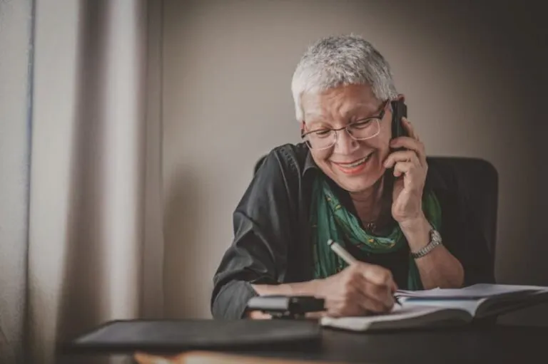 A senior woman is on a cell phone call while writing notes in a notebook