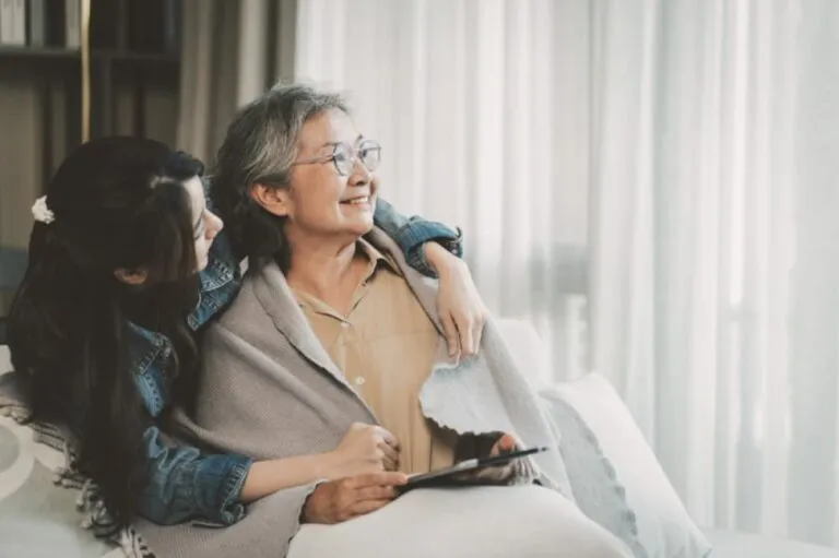 A senior woman lounging on a sofa with her daughter hugging her gently around the neck
