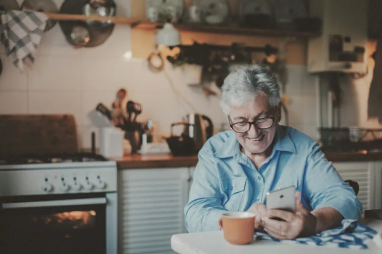 Older adult sitting at kitchen table smiling at phone.